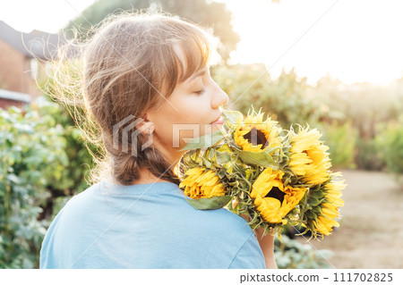 Dreaming young female farmer woman with closed eyes holding and sniffing a sunflowers bouquet on the green garden background in sunset light. Enjoy the moment. Rural, Cottage core lifestyle 111702825