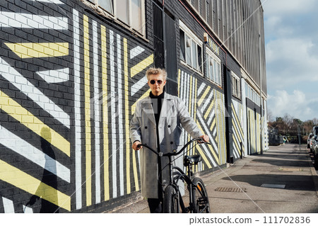 Stylish male in checked coat and sunglasses goes with his retro bicycle by painted urban wall in sunny day. Neutral carbon footprint transportation. Green eco friendly mobility sustainable transport 111702836