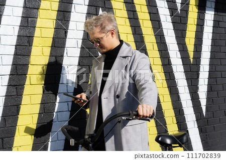 Stylish male in coat and sunglasses using phone while walking with retro bicycle by painted urban wall in sunny day. Neutral carbon footprint transportation. Ecofriendly mobility sustainable transport Stylish male in coat and sunglasses using phone while walking with retro bicycle by painted urban wall in sunny day. Neutral carbon footprint transportation. Ecofriendly mobility sustainable transport 111702839