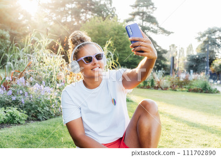 Young smiling lesbian lgbtq woman using mobile phone at park, having video chat with friends, doing online shopping, creating content for followers. Social Diversity acceptance, feel free lifestyle. 111702890