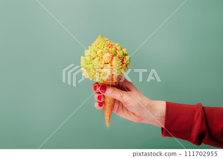 Female hand holding Waffle cone with Romanesco cabbage with dripping fruit sauce as vegetarian dessert on pastel green background. Plant based Ice cream. Creative healthy vegetarian and vegan food Female hand holding Waffle cone with Romanesco cabbage with dripping fruit sauce as vegetarian dessert on pastel green background. Plant based Ice cream. Creative healthy vegetarian and vegan food 111702955