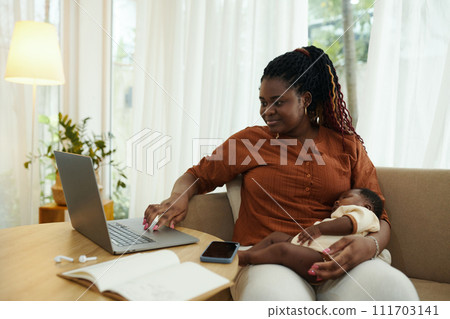 Smiling Black businesswoman sitting on couch with sleeping baby on laps and answering emails 111703141