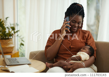 Portrait of Black woman sitting with baby on laps and taking on phone Portrait of Black woman sitting with baby on laps and taking on phone 111703143