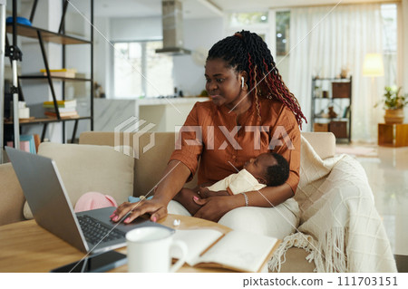 Black woman sitting on couch with newborn baby on laptop when having online meeting 111703151