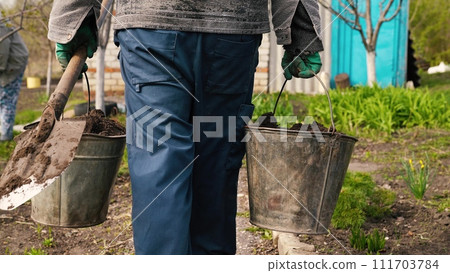 American farm, farmer carries two buckets earth, gardening business, caring plants, trees, farmer works garden, agricultural worker, man carries bucket earth, rear view, road earth field, going work 111703784