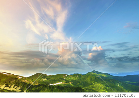 Wide panorama of lit by morning sun green valley, hills covered with forest and distant misty mountains with patches of snow under cloudy windy sky. Beauty of nature, tourism and traveling concept. Wide panorama of lit by morning sun green valley, hills covered with forest and distant misty mountains with patches of snow under cloudy windy sky. Beauty of nature, tourism and traveling concept. 111705103