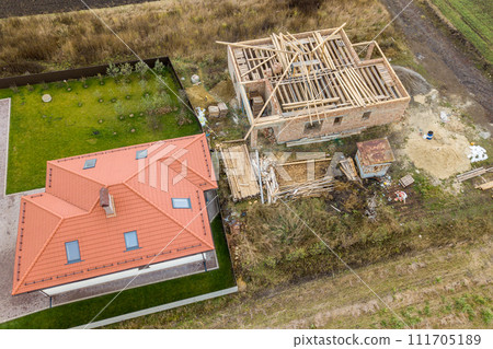 Top down aerial view of two private houses, one under construction with wooden roofing frame and another finished with red tiled roof. 111705189