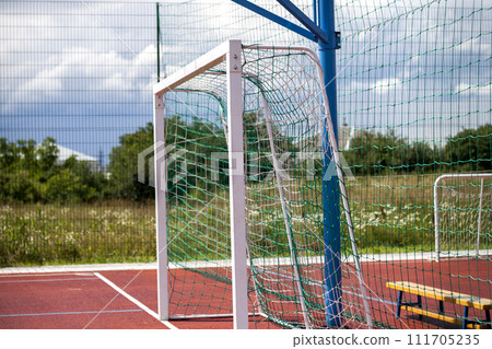 Soccer or football field playground with bright red soft rubber flooring, big gate, empty bench and protective net fence in rural countryside on blue sky, green grass and trees background. Soccer or football field playground with bright red soft rubber flooring, big gate, empty bench and protective net fence in rural countryside on blue sky, green grass and trees background. 111705235