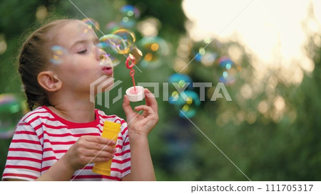 Spending time in park little girl finds joy in simple pleasure of blowing soap bubbles. Happy girl entertains by blowing bubbles in summer city park. Girl blows soap bubbles slow motion 111705317