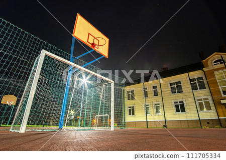 Outdoors mini football and basketball court with ball gate and basket surrounded with high protective fence brightly illuminated with spotlight lamps at night. 111705334