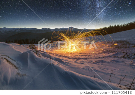 Light painting art. Spinning steel wool in abstract circle, firework of bright yellow glowing sparkles on snowy white valley on woody mountain ridge and blue night starry sky copy space background. 111705418