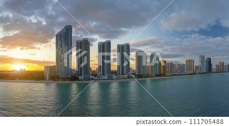High angle view of Sunny Isles Beach city at sunset with expensive highrise hotels and condo buildings over beachfront on Atlantic shore. American tourism infrastructure in coastal southern Florida High angle view of Sunny Isles Beach city at sunset with expensive highrise hotels and condo buildings over beachfront on Atlantic shore. American tourism infrastructure in coastal southern Florida 111705488
