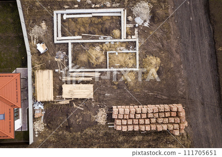 Concrete foundation for basement of future house, stacks of bricks and building timber logs for construction on sunny summer day, aerial view. Concrete foundation for basement of future house, stacks of bricks and building timber logs for construction on sunny summer day, aerial view. 111705651