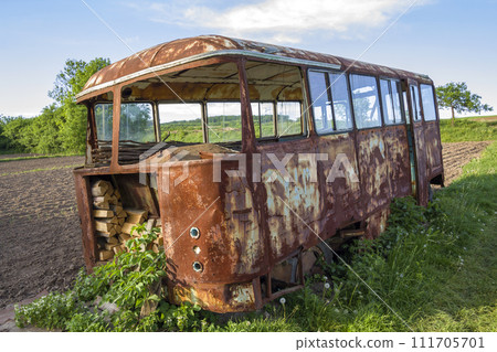Close-up of old forsaken passenger bus with broken windows rusting in high green weedy grass on edge of plowed brown field on bright spring day under blue morning sky. 111705701