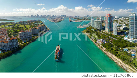 Aerial view of large container ship entering in Miami harbor main channel near South Beach. Luxurious hotels and apartment buildings on waterfront and high skyscrapers of downtown district in distance Aerial view of large container ship entering in Miami harbor main channel near South Beach. Luxurious hotels and apartment buildings on waterfront and high skyscrapers of downtown district in distance 111705963