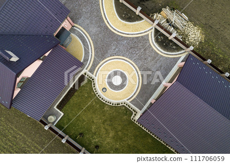 Aerial top view of house shingle roof on background of green lawn and colorful paved yard with geometrical abstract pattern. Roofing, repair and renovation work. Aerial top view of house shingle roof on background of green lawn and colorful paved yard with geometrical abstract pattern. Roofing, repair and renovation work. 111706059