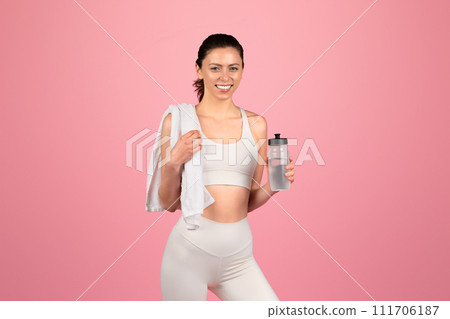 A happy, athletic woman in a white sports bra and leggings holds a water bottle and a towel over her shoulder A happy, athletic woman in a white sports bra and leggings holds a water bottle and a towel over her shoulder 111706187