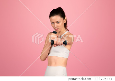 Focused young woman in white sportswear engaging in a strength training session with a black dumbbell 111706275