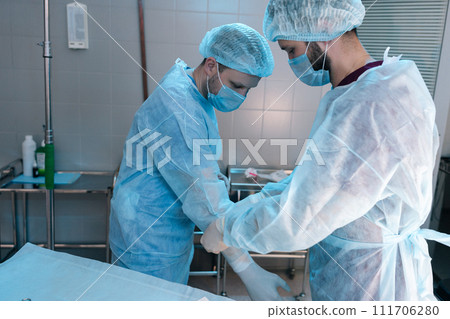 Assistant male helping surgeon putting on latex gloves before operation. Team of surgeons in operating room with light modern interior preparing for surgery. Concept of surgery and emergency. 111706280