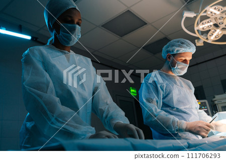 Shooting from below of male surgeon assisted by African female nurse in gown, mask and protective gloves using surgical instruments to operating patient in operation room. Concept of healthcare. Shooting from below of male surgeon assisted by African female nurse in gown, mask and protective gloves using surgical instruments to operating patient in operation room. Concept of healthcare. 111706293