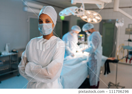 Portrait of confident African-American female doctor standing posing with crossed arms looking at camera in operating theater. Diverse team of surgeons performing operation on background. 111706296