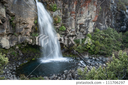 Waterfall bursting from rocky clif into small pool surrounded by exotic foliage, New Zealand 111706297
