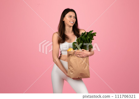 Smiling young woman in athletic wear holding a paper grocery bag full of fresh vegetables and fruits Smiling young woman in athletic wear holding a paper grocery bag full of fresh vegetables and fruits 111706321
