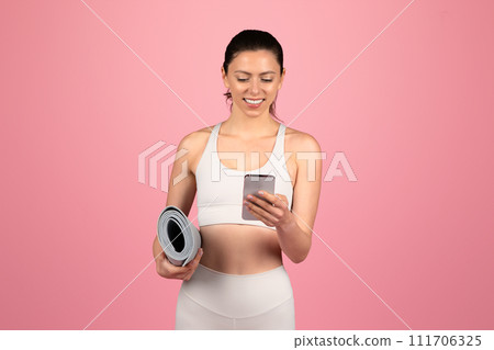 Fitness-focused woman in a white sports bra and leggings holding a yoga mat and using a smartphone 111706325