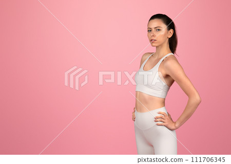Determined young woman in white fitness attire standing confidently with hands on hips 111706345