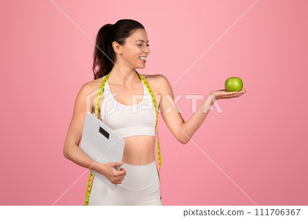 Smiling fitness-oriented woman with a measuring tape over her shoulder holding a scale 111706367