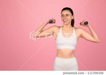 Energetic woman in sportswear flexing her biceps with dumbbells, showing off her strength 111706398