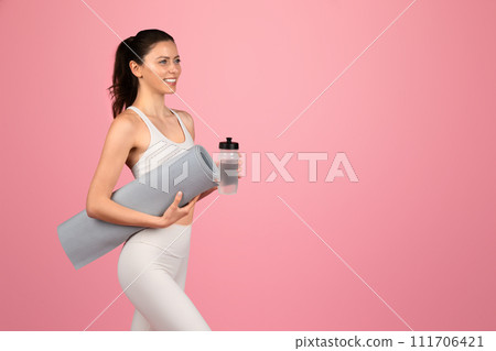 A smiling woman in a white tank top and leggings carrying a yoga mat and water bottle 111706421