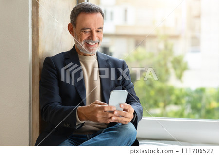 Smiling older man using a smartphone by the window Smiling older man using a smartphone by the window 111706522