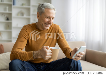 Smiling old man sitting on couch at home, holding phone 111706882