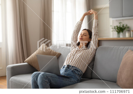 Relaxed young woman stretching on sofa with laptop Relaxed young woman stretching on sofa with laptop 111706971