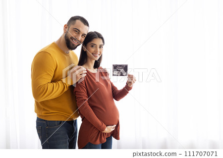 Pregnant Young Couple Showing Baby Sonogram While Standing Near Window At Home Pregnant Young Couple Showing Baby Sonogram While Standing Near Window At Home 111707071