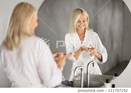 Beautiful senior woman using moisturising lotion while standing near mirror in bathroom Beautiful senior woman using moisturising lotion while standing near mirror in bathroom 111707292