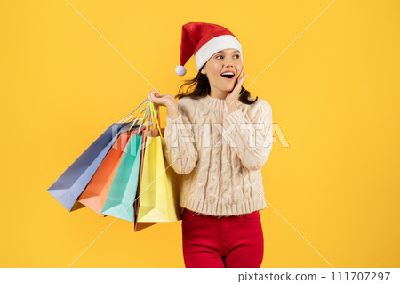 lady shopper in Santa hat holding shopping bags, yellow backdrop 111707297