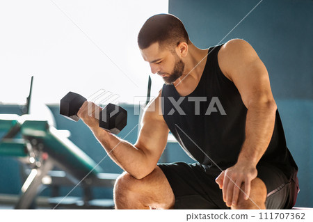Young Male Athlete Training With Dumbbell At Gym, Making Seated Biceps Curl Young Male Athlete Training With Dumbbell At Gym, Making Seated Biceps Curl 111707362