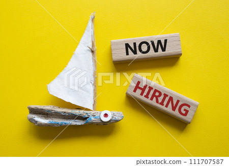 Now Hiring symbol. Concept word Now Hiring on wooden blocks. Beautiful yellow background with boat. Business and Now Hiring concept. Copy space Now Hiring symbol. Concept word Now Hiring on wooden blocks. Beautiful yellow background with boat. Business and Now Hiring concept. Copy space 111707587