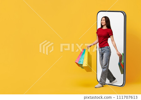 Cheerful young asian lady with purchases walking from huge phone Cheerful young asian lady with purchases walking from huge phone 111707613