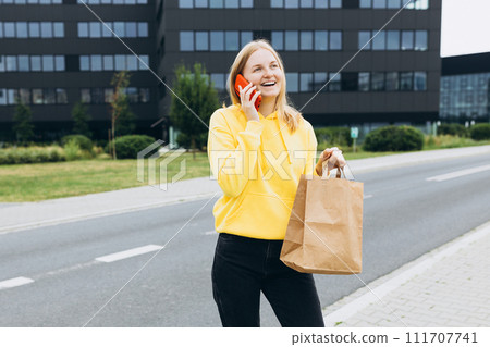 30s happy woman holding a paper bag in hands and calling on phone on city street. Technology, communication and people concept 111707741