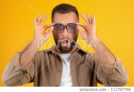 Surprised young caucasian man with beard in casual with open mouth, takes off his glasses 111707746