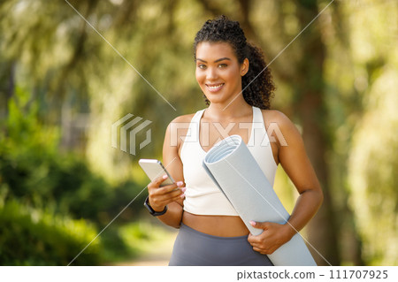 Young fitness woman holds yoga mat browsing smartphone outdoors Young fitness woman holds yoga mat browsing smartphone outdoors 111707925