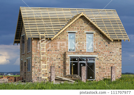 Two-story residential house with plastic windows, wide door and wooden frame for roof under construction. High wooden ladder at brick walls and brick columns for fence on dark blue sky background. Two-story residential house with plastic windows, wide door and wooden frame for roof under construction. High wooden ladder at brick walls and brick columns for fence on dark blue sky background. 111707977