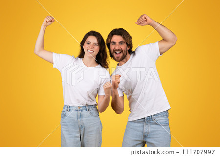 Enthusiastic young couple in white t-shirts and jeans cheerfully raising fists in a victorious gesture 111707997