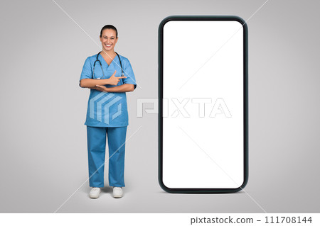 Cheerful european woman nurse, surgeon in blue uniform point at big phone with empty screen, isolated on gray background Cheerful european woman nurse, surgeon in blue uniform point at big phone with empty screen, isolated on gray background 111708144