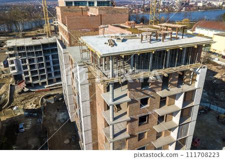 Apartment or office tall building under construction, top view. Brick walls, scaffolding and concrete support pillars. Tower crane on bright blue sky copy space background. Drone aerial photography. Apartment or office tall building under construction, top view. Brick walls, scaffolding and concrete support pillars. Tower crane on bright blue sky copy space background. Drone aerial photography. 111708223