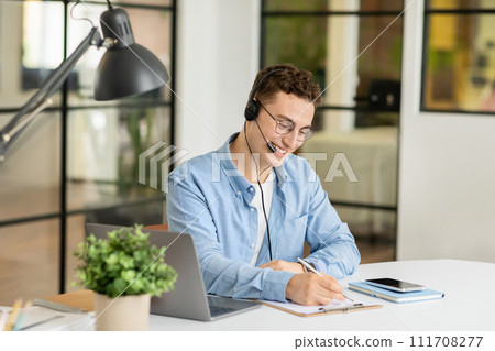Positive handsome millennial european guy manager in headphones with laptop at table Positive handsome millennial european guy manager in headphones with laptop at table 111708277