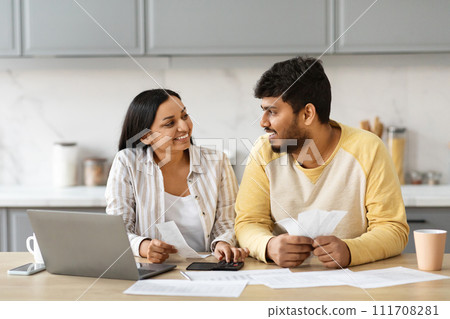 Smiling indian couple with laptop in kitchen calculating bills 111708281